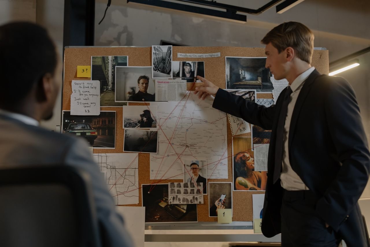 Services Two detectives discussing suspects and evidence on a cork board in an office setting.